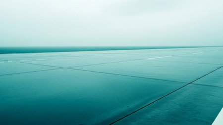 presents a wide, wet asphalt road that recedes into a hazy, misty horizon. The surface of the road is slick with water, reflecting the pale, overcast sky in muted teal and grey tones. White lines mark the lanes, providing a sense of direction and scale. The atmosphere is serene and slightly melancholic, with the mist obscuring the distant landscape and creating a sense of vastness and solitude. The composition is minimalist, focusing on the clean lines of the road and the atmospheric of the...の素材