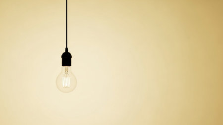 A solitary incandescent light bulb hangs suspended by a thin black cord against a plain, pale yellow or beige background. The glass bulb is clear, revealing the intricate filament within. The lighting is soft, subtly illuminating the bulb and creating a gentle glow. The composition is minimalist and clean, with ample negative space, making the light bulb the sole focal point. The evokes a sense of simplicity, ideas, or energy.の素材