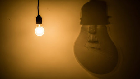 A close-up shot of a single, old-fashioned incandescent light bulb hanging from a black wire against a warm, dark background. The bulb is brightly illuminated, emitting a soft, yellowish-orange glow. A distinct, enlarged shadow of the light bulb is cast on the wall behind it, creating a strong contrast and a sense of depth. The focuses on the simple yet essential of light.の素材