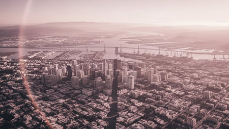 An expansive aerial perspective captures a city waking up at dawn. The soft morning light casts a gentle glow over a dense urban landscape filled with numerous buildings and skyscrapers. Residential areas with smaller structures fill the foreground, transitioning to a more industrial and zone with a large port and harbor. Bridges span across waterways, connecting different parts of the city. The distant horizon is softened by atmospheric haze, creating a serene and vast panorama.の素材