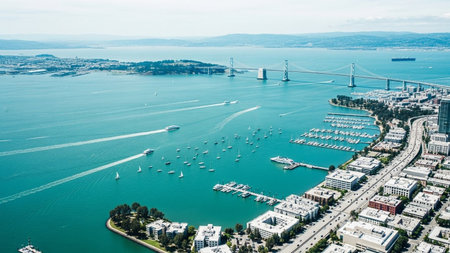 An elevated, wide-angle aerial view of the San Francisco Bay on a bright, sunny day. The expansive turquoise water is dotted with numerous sailboats and yachts, many docked in a bustling marina. A prominent suspension bridge, likely the Bay Bridge, stretches across the water, connecting the city to the distant land. The city's urban landscape with its buildings and coastline curves along the edge of the bay. The scene is vibrant and showcases a blend of urban development and recreational...の素材