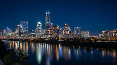 The Austin, Texas skyline is dramatically illuminated against a deep blue night sky. Tall skyscrapers and modern buildings are lit up, casting a vibrant glow. The city lights are beautifully reflected in the dark, still waters of the Colorado River in the foreground. A bridge spans the river, also illuminated. The overall scene is serene and visually striking, showcasing the urban beauty of the city at night with its shimmering reflections.の素材