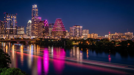 The Austin, Texas skyline is captured at night, showcasing a vibrant display of illuminated skyscrapers and modern architecture. The Colorado River flows in the foreground, reflecting the colorful lights of the city and creating streaks of light from passing traffic on the bridge. Lush green trees line the riverbanks, contrasting with the urban glow. The deep blue of the night sky provides a dramatic backdrop to the glowing city lights, with prominent pink and purple hues emanating from some...の素材