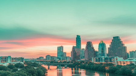 The Austin skyline is captured at sunset with a dramatic sky painted in vibrant hues of pink, orange, and teal. The Colorado River flows through the foreground, reflecting the colorful sky and the city's modern architecture. Lush green trees line the riverbanks, contrasting with the urban landscape. The scene is tranquil and showcasing the city's blend of nature and urban development.の素材