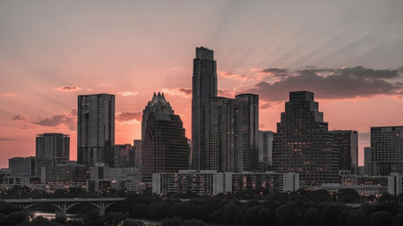 A panoramic view of the Austin, Texas skyline at sunset. The sky is painted with vibrant hues of orange, pink, and purple, with dramatic sun rays piercing through scattered clouds. Tall modern skyscrapers dominate the cityscape, their silhouettes softened by the fading light. The urban landscape stretches towards the horizon, showcasing the dynamic energy of the city.の素材