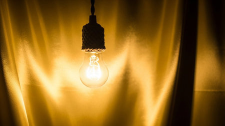 A single incandescent light bulb hangs from a cord, emitting a warm, yellow glow. The bulb is illuminated, revealing its internal filament. Behind it, a textured curtain creates a soft, diffused background that enhances the warm ambiance of the light. The evokes a sense of creativity, inspiration, or a cozy, inviting atmosphere.の素材