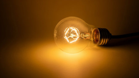 A close-up studio shot of a classic incandescent light bulb lying on its side. The bulb is illuminated, casting a warm, golden-yellow glow. The intricate, coiled filament inside the glass bulb is clearly visible, glowing brightly and creating a visual. The background is a soft, warm gradient of yellow and orange, enhancing the cozy and atmospheric feel of the focus is sharp on the bulb and its filament, highlighting the delicate structure and the emitted light.の素材
