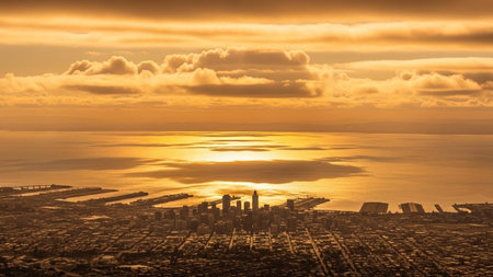 An aerial view captures a sprawling cityscape bathed in the warm glow of a golden sunset. The sun's rays reflect brilliantly on the calm ocean waters, creating a shimmering path of light. Distant industrial ports and harbors are visible along the coastline, hinting at the city's economic activity. Wispy clouds drift across the sky, adding texture to the atmospheric scene. The overall mood is one of serene beauty and expansive tranquility.の素材