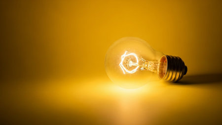 A single incandescent light bulb lies on its side against a warm, gradient yellow background. The filament inside the bulb is brightly glowing, casting a soft, diffused light that illuminates the surrounding area and creates a subtle shadow. The has a warm, inviting, and slightly nostalgic feel due to the color palette and the vintage nature of the bulb. The focus is on the light source and its radiant glow, evoking a sense of warmth, energy, and the concept of an idea.の素材