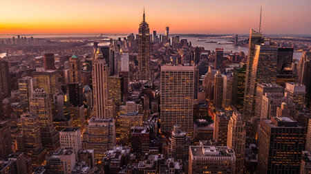 An aerial panorama of the New York City skyline at sunset. The city stretches out to the horizon, a dense collection of diverse skyscrapers illuminated by the warm, golden light of the setting sun. The sky is a gradient of orange, pink, and purple hues, casting a magical glow over the iconic architecture. captures the immense scale and vibrant energy of one of the world's most famous cities, travel, urban planning, or business-related themes.の素材