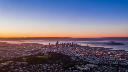 A aerial view showcases the San Francisco cityscape and its surrounding bay during a vibrant sunrise. The sky transitions from deep blue to soft oranges and pinks, casting a warm, golden light over the urban landscape. The iconic Golden Gate Bridge is visible in the distance, spanning the bay. The city's dense architecture, rolling hills, and the vast expanse of water create a visual composition. This panoramic shot captures the serene beauty of the city waking up, with clear skies and the...の素材