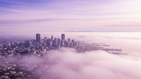 This aerial view showcases the San Francisco skyline partially obscured by a dense, ethereal blanket of fog, tinged with soft purple and pink hues from the dawn light. Tall skyscrapers rise above the swirling mist, creating a dreamlike and serene urban landscape. The fog rolls in from the bay, enveloping the city in a soft, atmospheric embrace. The scene is characterized by its pastel color palette and a sense of peaceful isolation.の素材