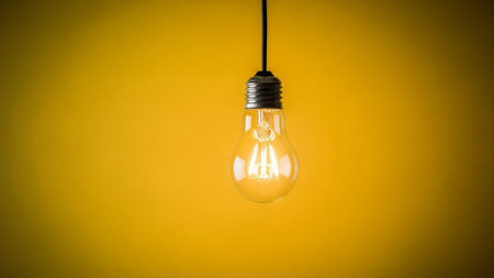 A single, clear glass incandescent light bulb hangs suspended by a black cord against a gradient yellow background that darkens towards the edges. The filament inside the bulb is visible and appears to be glowing faintly, suggesting warmth and illumination. The composition is minimalist and focuses on the iconic shape of the bulb, symbolizing ideas and inspiration.の素材