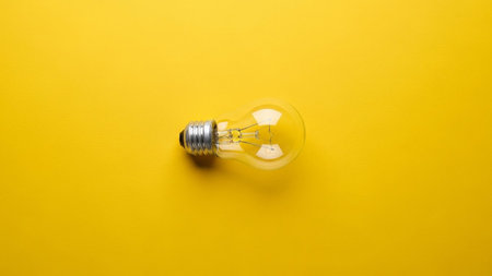 A single, clear incandescent light bulb lies horizontally on a stark, bright yellow background. The bulb's glass is transparent, revealing the intricate filament within, and it has a standard silver metal screw base. The composition is extremely minimalist and clean, with the vibrant yellow providing a strong contrast to the neutral tones of the light bulb. is representing concepts such as ideas, innovation, inspiration, or energy due to the iconic nature of the light bulb. The ample plain...の素材