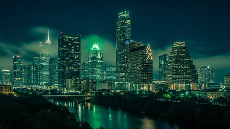 The Austin, Texas skyline is captured at night, showcasing a dense collection of illuminated skyscrapers. The city lights twinkle against the dark sky, with a river flowing in the foreground, mirroring the urban glow. The architecture is modern and varied, with some buildings featuring distinctive lighting. The overall impression is one of a vibrant and bustling metropolitan area after dark.の素材