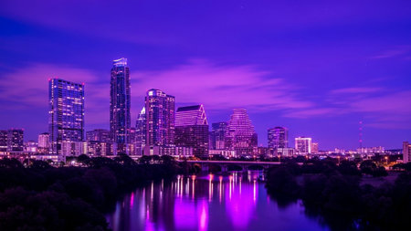 A cityscape bathed in vibrant purple and violet hues. Modern skyscrapers with illuminated windows and neon accents pierce the deep purple twilight sky. The city's glow is beautifully mirrored in the calm, dark waters of a river flowing through the foreground, creating a reflection of the urban landscape. Wispy clouds add texture to the sky, enhancing the atmospheric and serene mood of the scene.の素材