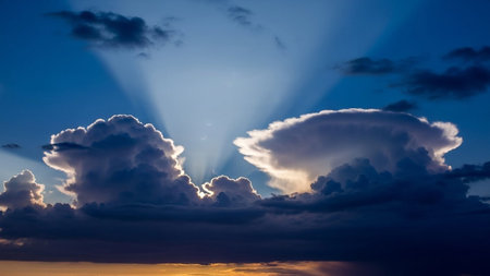 Dramatic crepuscular rays emanate from behind dark cumulonimbus clouds against a deep blue sky with hints of orange...の素材