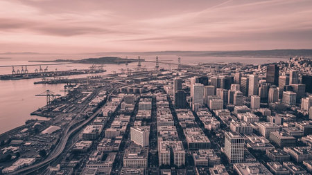 An expansive aerial perspective captures a vast coastal city bathed in the soft, muted tones of dusk. The urban landscape stretches towards the horizon, characterized by a dense arrangement of modern buildings and skyscrapers. A prominent bridge spans a body of water, connecting different parts of the city. Industrial areas with docks and cranes are visible along the coastline, hinting at the city's economic activity. The sky above is filled with subtle clouds, casting a pinkish hue over the...の素材