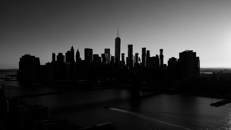 A striking black and white aerial view captures the dramatic silhouette of a vast city skyline against a dark, clear sky at dusk. Numerous skyscrapers and buildings of varying heights form a dense urban landscape. A body of water, likely a river or bay, lies in the foreground, with a small boat leaving a visible wake. The contrast and lack of color create a powerful and moody atmosphere, emphasizing the imposing scale of the metropolis.の素材