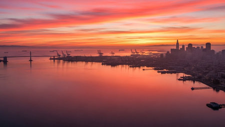 A aerial view captures the San Francisco cityscape and its bay during a vibrant sunset. The sky is a dramatic canvas of fiery oranges and soft pinks, with wisps of clouds reflecting on the calm water. Silhouetted against the colorful sky are the city's iconic skyline, the distant Oakland Bay Bridge, and a bustling harbor filled with cranes and ships. The scene is both industrial and showcasing the city's unique blend of urban life and natural splendor.の素材
