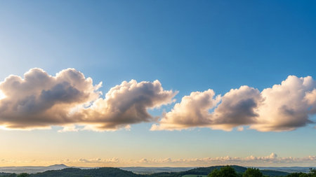 Golden sunlight illuminates fluffy cumulus clouds against a vibrant blue sky, casting a warm glow over distant green...の素材