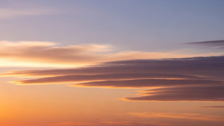 Streaks showing wispy lenticular clouds illuminated by the warm orange and purple hues of a setting sun in a clear...の素材