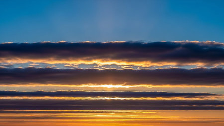 A dramatic sunset sky with layered dark clouds illuminated by golden sunlight creating a vibrant horizon.の素材