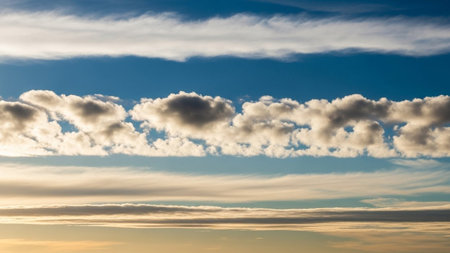 A wide sky distinct horizontal bands of white and grey clouds against a bright blue sky, touched by soft golden light.の素材