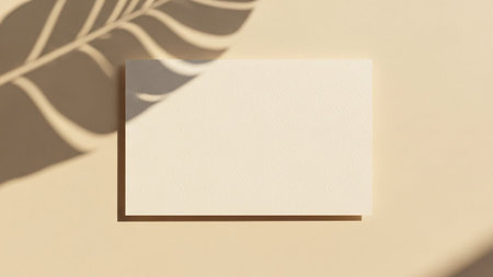 A clean, minimalist composition featuring a blank beige rectangular card placed on a matching beige surface. A prominent shadow of a large leaf is cast across the left side of the card and background, creating a natural and organic feel. Soft, warm light illuminates the scene, highlighting the subtle texture of the paper and casting gentle shadows. is mockups, stationery or conveying a sense of calm and simplicity.の素材