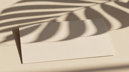 A minimalist composition featuring a blank, beige rectangular card placed on a neutral, light-colored surface. The card is bathed in soft sunlight, casting a distinct shadow from a tropical leaf, possibly a palm or fern, onto its surface and the background. The shadow creates an organic, dappled pattern, adding a touch of nature and visual interest to the clean aesthetic. The overall impression is serene and uncluttered, offering ample copy space for or branding purposes. The interplay of...の素材