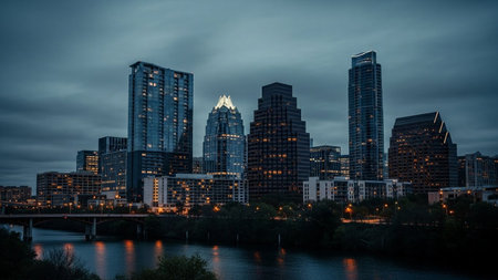A panoramic view of a modern city skyline as dusk settles. Tall skyscrapers with illuminated windows pierce the darkening sky, casting a warm glow. A calm river flows in the foreground, reflecting the city lights and the moody twilight atmosphere. Trees line the riverbank, adding a touch of nature to the urban landscape. The scene evokes a sense of urban grandeur and tranquility.の素材