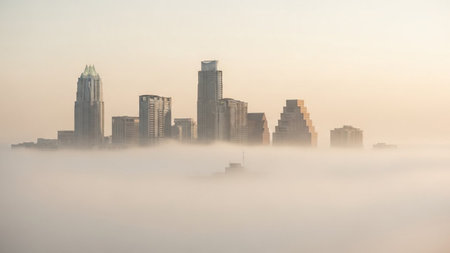 A serene cityscape is partially obscured by a thick blanket of fog during sunrise. The soft golden light of dawn gently illuminates the tops of the skyscrapers, creating an ethereal and dreamlike atmosphere. The mist creates a sense of depth and mystery, with only the upper portions of the buildings visible above the hazy layer. The sky is a pale, diffused gradient of warm tones, contributing to the tranquil and peaceful mood of the scene.の素材