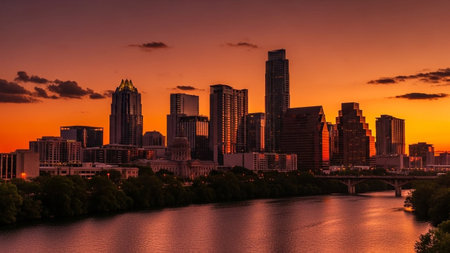 The Austin, Texas skyline is dramatically illuminated by a vibrant orange sunset. Tall skyscrapers and modern architecture rise against a colorful sky, with clouds adding texture and depth. The Colorado River flows in the foreground, perfectly reflecting the city lights and the warm hues of the twilight sky. A bridge spans the river, connecting the urban landscape. captures the beauty and energy of the city at dusk, offering a scenic and atmospheric view.の素材