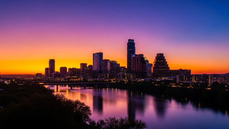 The Austin, Texas skyline is captured at dusk, bathed in the vibrant colors of a twilight sky transitioning from deep purple to bright orange. The city's modern skyscrapers are silhouetted against this colorful backdrop. The calm waters of the Colorado River in the foreground perfectly reflect the illuminated buildings and the dramatic sky, creating a symmetrical and serene scene. Trees along the riverbank add a touch of natural darkness to the foreground.の素材