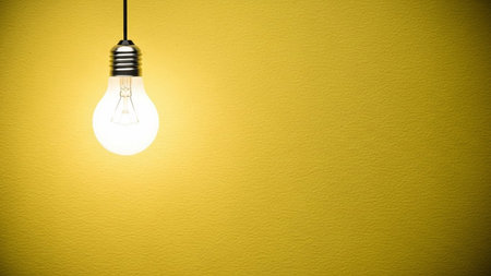 A single incandescent light bulb hangs from a wire, emitting a warm, bright glow. The bulb is illuminated against a textured yellow wall, creating a soft contrast. The focus is on the light source and its immediate surroundings, with ample negative space on the right side of the frame. The evokes a sense of simplicity, ideas, and illumination.の素材