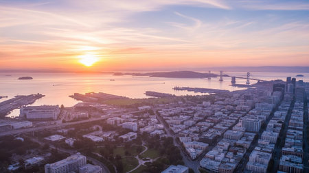 An aerial view captures the sprawling San Francisco skyline at sunset. The sun, a bright orb on the horizon, casts a warm, orange and pink glow across the sky and the calm waters of the bay. The iconic Bay Bridge stretches across the water, its structure silhouetted against the vibrant sky. Numerous buildings of varying heights form the dense cityscape, with industrial areas and a park visible closer to the foreground. The scene is peaceful and visually highlighting the beauty of the city at...の素材
