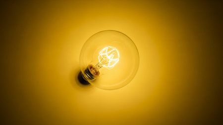 A close-up, top-down view of a single, vintage-style incandescent light bulb. The filament inside the bulb is clearly visible and glowing with a warm, bright yellow light. The bulb is set against a solid, warm yellow background, creating a soft, diffused light that emanates from the focuses on the detail of the filament and the overall warm illumination.の素材