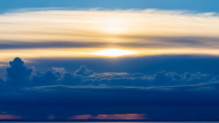A dramatic sunset sky with a bright golden sun peeking through layers of dark blue and white clouds over a calm,...の素材