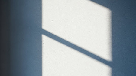 A sharp, geometric shadow is cast diagonally across a textured white wall, creating a minimalist and serene scene.の素材