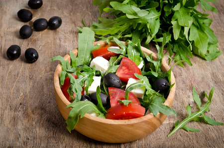 Salad with tomatoes and olives on wooden background. a bowl of salad and a bunch of arugula with olives behindの写真素材