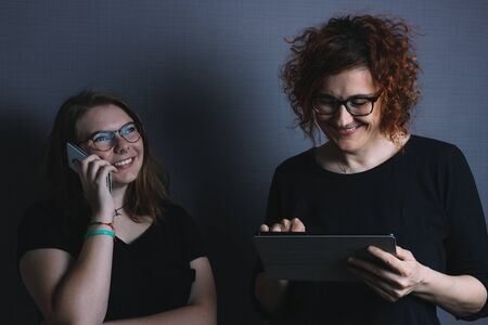 young blonde girl talking on the phone and redhead woman with tablet, happy, smiling, wearing black t-shirtの写真素材