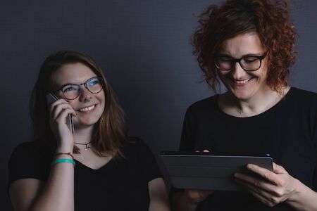 young blonde girl talking on the phone and redhead woman with tablet, happy, smiling, wearing black t-shirtの写真素材