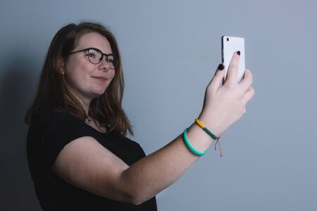 young blonde girl with glasses interacting with a smartphone, on a gray background, taking a selfieの写真素材