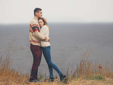 Young attractive couple in knitted sweaters standing on a cliff at sea shore and huggingの写真素材