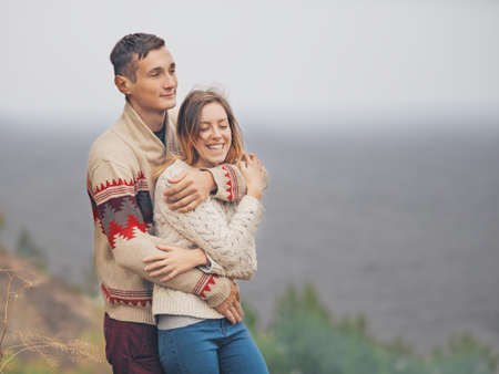 Young attractive couple in knitted sweaters standing on a cliff at sea shore and huggingの写真素材