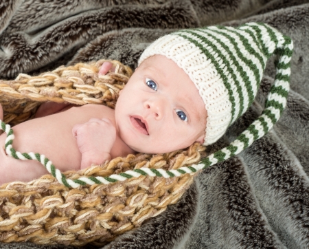Adorable blue eyed newborn in a knit basket with a green and white knit cap on a fur blanketの写真素材