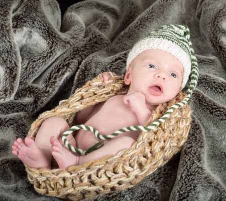 cute baby in a knit basket wearing a green and white knit hat on a fur blanketの写真素材
