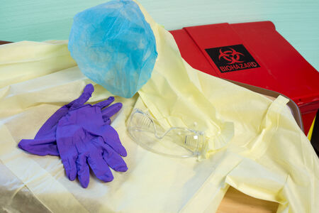 Disposable hospital gown, gloves, hair cover and goggles on a table next to a biohazard trash can against a light green wallの写真素材