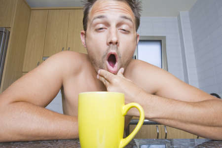 Portrait of a yawning man in the kitchen with a colorful yellow mug of coffee.の写真素材