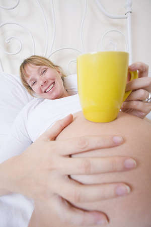 Pregnant woman in bed with a yellow mug on her bumpの写真素材